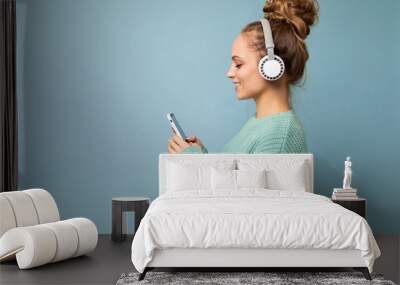 Photo of beautiful joyful smiling young woman wearing stylish casual clothes isolated over background wall holding and using mobile phone wearing white bluetooth headphones listening to music and Wall mural