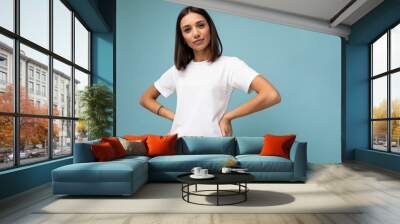 Photo of positive young beautiful brunette woman with sincere emotions wearing casual white t-shirt for mockup isolated over blue background with copy space Wall mural