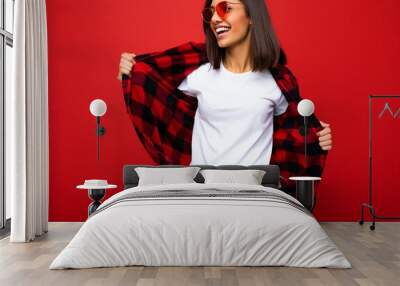 Photo of young beautiful happy smiling brunette woman wearing trendy white t-shirt with empty space for mock up and red check shirt and red sunglasses. Sexy carefree female person posing isolated near Wall mural