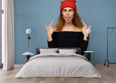 Photo of young positive beautiful brunette curly woman with sincere emotions wearing black crop top and red hat isolated on blue background with copy space and grimacing Wall mural