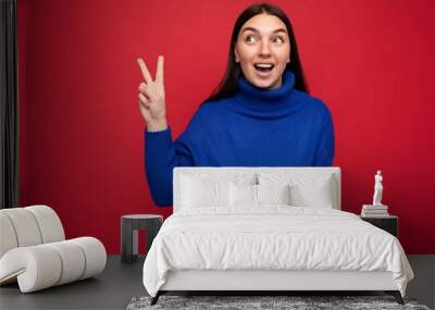 Photo of young positive happy smiling beautiful woman with sincere emotions wearing stylish clothes isolated over background with copy space and showing peace gesture with two fingers Wall mural