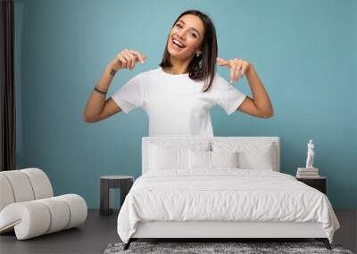 Portrait of positive happy smiling young beautiful brunette woman with sincere emotions wearing casual white t-shirt for mockup isolated over blue background with copy space and pointing at empty Wall mural