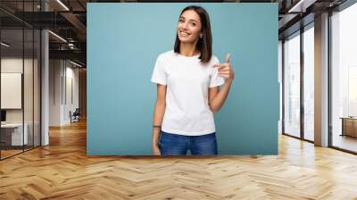 Portrait of young beautiful happy smiling brunette woman wearing trendy white t-shirt with empty space for mock up. Sexy carefree female person posing isolated near blue wall in studio with free space Wall mural