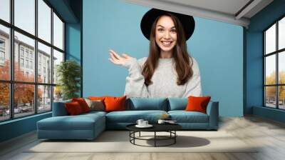 Portrait of young positive happy smiling nice winsome beautiful brunette woman with sincere emotions wearing stylish grey pullover and black hat isolated over blue background with copy space Wall mural