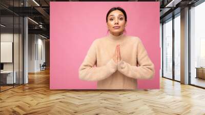 Portrait of young serious beautiful brunette woman with sincere emotions wearing casual beige jersey isolated over pink background with free space and keeping hands together asking for forgiveness Wall mural