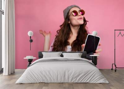 Positive attractive young brunette female person wearing stylish pink shirt grey hat and colourful sunglasses isolated over pink background holding in hand and showing mobile phone with empty display Wall mural