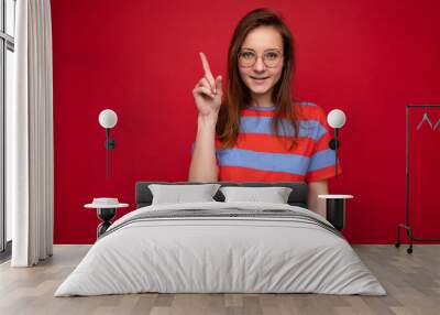 Positive happy young attractive brunette woman with sincere emotions wearing casual striped t-shirt and optical glasses isolated over red background with empty space and showing one finger Wall mural