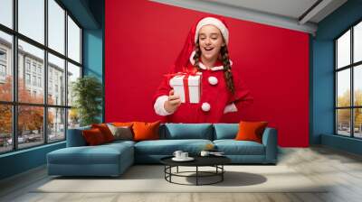 Shot of attractive happy smiling brunette girl isolated over red background wall wearing Santa Claus suit holding white gift box with red ribbon and looking at surprise Wall mural