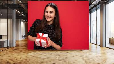 Shot of charming happy smiling brunette girl isolated over red background wall wearing black blouse holding white gift box with red ribbon and looking at camera Wall mural