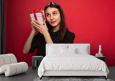 Shot of charming happy thoughtful brunette girl isolated over red background wall wearing black blouse holding white gift box with red ribbon and looking up Wall mural