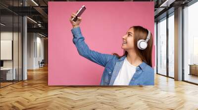 Side-profile Photo of attractive positive smiling young dark blonde woman wearing blue jean stylish shirt and casual white t-shirt isolated over pink background wearing white bluetooth wireless Wall mural