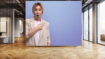 surprised blond business woman in office jacket pointing finger towards advertisement on studio background Wall mural