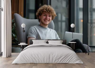 Young caucasian man with curly hair comfortably sitting in armchair working or using laptop with space for text
 Wall mural