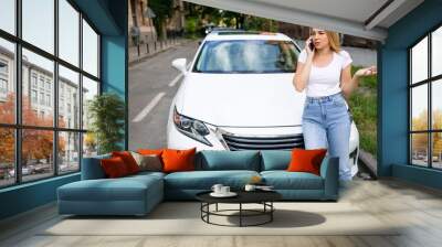 Young woman standing near broken car. Girl using smartphone calling for help. Attractive girl interrupted the trip because of a breakdown Wall mural