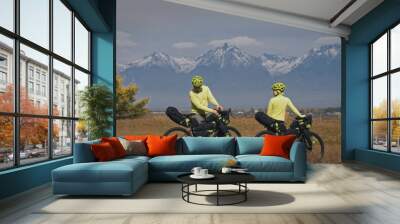 The man and woman travel on mixed terrain cycle touring with bikepacking. The two people journey with bicycle bags. Sport bikepacking, bike, sportswear in green black colors. Mountain snow capped. Wall mural