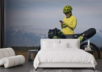 The woman travel on mixed terrain cycle touring with bikepacking. The traveler journey with bicycle bags. Sportswear in green black colors. Mountain snow capped. Make a selfie smartphone Wall mural