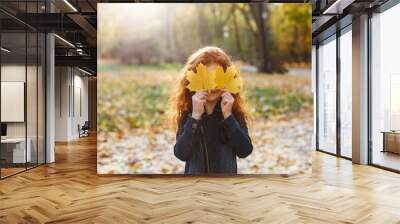 Autumn vibes, child portrait. Charming and red hair little girl looks happy walking and playing on the fallen leaves in autumn park full of evening sunshine Wall mural