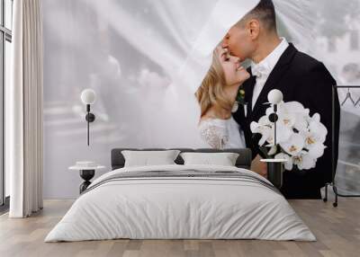 Groom in black tuxedo hugs tender stunning bride while they stand on the street of old European town Wall mural