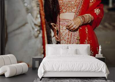 Indian groom dressed in white Sherwani and red hat with stunning bride in red lehenga stand and hold each hands walking outside Wall mural