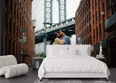 Love story in New York. Gorgeous couple of American man with beard and tender Eastern woman hug each other before the cityscape of Brooklyn bridge somewhere in New York Wall mural