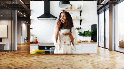 Smiled attractive girl is eating cutted fruits on white modern kitchen dressed in nightwear with messy loose hair Wall mural