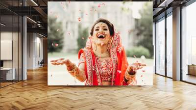 Stunning Indian bride dressed in Hindu traditional wedding clothes lehenga embroidered with gold and a veil smiles tender posing outside with golden accessories under the rain of petals Wall mural