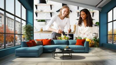 Two women of different nationalities are smiling and cooking a salad in the kitchen Wall mural