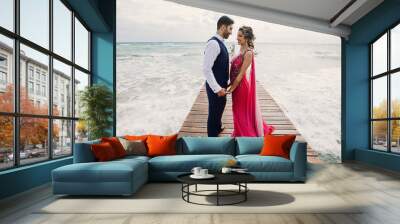 Wind blows bride's pink sari while she stands with Hindu groom on a wooden quay among foaming waves Wall mural