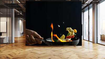 Tossing vegetables in a cooking pan with flames of fire around them suggesting flambe by covering them with spirit and set alight briefly. Black background and chef's hand holding the pan. Wall mural