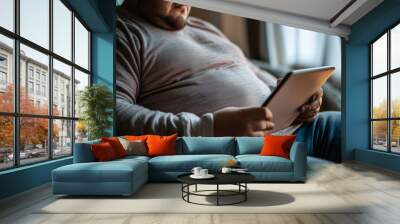 Very overweight individual browsing social media on a tablet, seated comfortably. A man is comfortably sitting on a couch while using a tablet device Wall mural