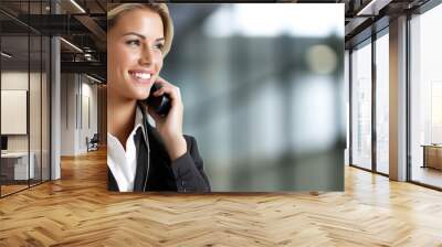 Professional woman in business attire smiling while talking on smartphone inside a modern office environment during daytime Wall mural