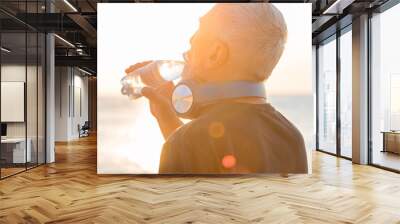 Old man athlete with gray beard and hair in headphones drinking water from a bottle on the background of the sea and dawn sky Wall mural