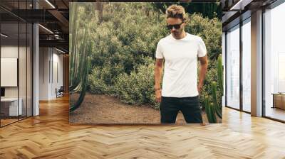 Young handsome bearded guy wearing a blank white t-shirt and sunglasses is standing in the garden background next to a big cactus plant. Horizontal mock up style Wall mural