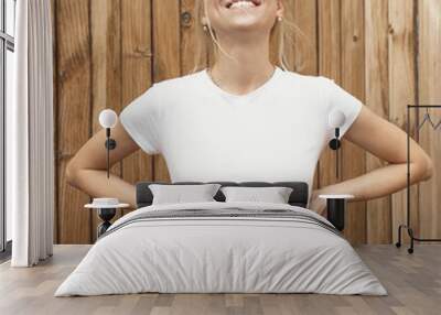 Young pretty girl is standing on a light wooden wall background. Woman is wearing a white empty t-shirt without logo which makes it being perfectly suitable for mock-up. Wall mural