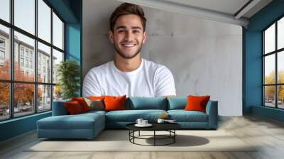 Cheerful young man with a bright smile, wearing a white T-shirt, arms folded, against a textured grey wall Wall mural