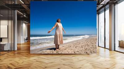 Middle aged long-haired woman in beige dress standing with her arms spread out on sandy beach by sea on beautiful sunny day. Energy and relax on beach. Front view Wall mural