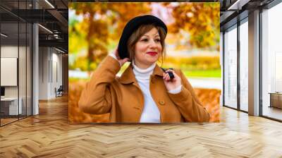 Portrait of beautiful brown-haired young woman wearing beige coat, cream turtleneck, black brimmed hat standing in city park on autumn day.	 Confidence. Wall mural