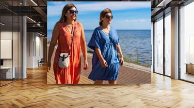 Summer vacation. Portrait of two pretty mid-adult women wearing flowing dresses walking on sidewalk by seaside resort on beautiful sunny day. Wall mural