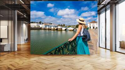 Summer vacation. Young woman with backpack in flowing clothes and hat standing by the river of the old city of Tavira Portugal and looking at roman bridge on a summer day. Visit the old town of Tavira Wall mural