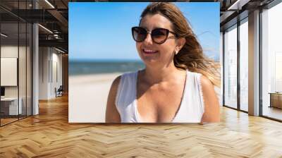 Summer. Portrait of young beautiful smiling woman with long blonde hair in light top and sunglasses against background of sandy beach and sea on sunny day. Energy, joy of life and youth Wall mural