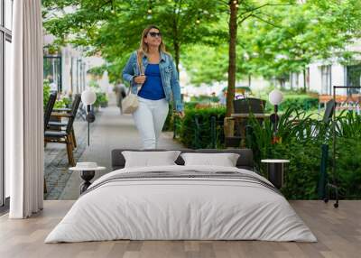 Young slim smiling woman in white pants and blue blouse and denim jacket walking down the street among gardens of cafes and restaurants in city center on sunny day. Everyday life in city Wall mural