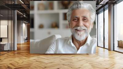 Close-up portrait of a smiling gray-haired senior man at home, sitting on a sofa in a white shirt and with a beard, smiling at the camera Wall mural