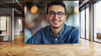 Close-up portrait of a young smiling man in glasses and a blue shirt sitting and working in the office and looking confidently at the camera Wall mural