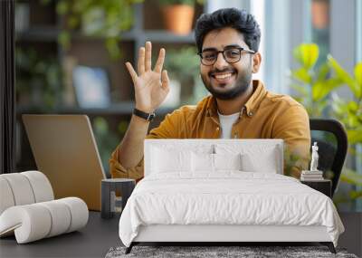 Happy young Indian man working in the office, sitting with a chair and enjoying success, looking at the laptop screen and showing a victory gesture with his hand Wall mural