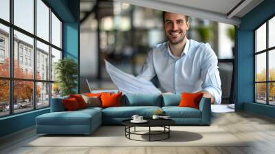 Portrait of a young businessman smiling at the camera, sitting at a desk in the office and working with documents and an invoice Wall mural