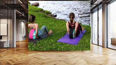 Back view two sportswomen doing yoga exercise with yoga wheel  near river in public park Wall mural