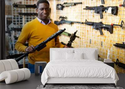 African-american man choosing assault rifle and shotgun in airsoft shop. Wall mural