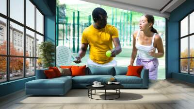 African american man instructor with a racket and a ball teaches a young woman to play padel tennis on an outdoor court Wall mural