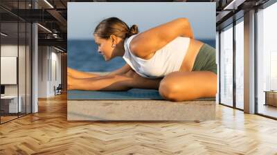 Against background of bright blue sea, slender of woman in sports clothes, hatha yoga lovers practicing oriental system of exercises. Woman performs Parivritta Janu Shirshasana Wall mural