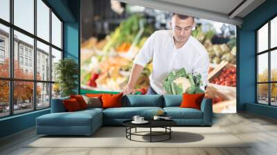 Careful young male customer buying fresh cauliflower in grocery store with large assortment Wall mural